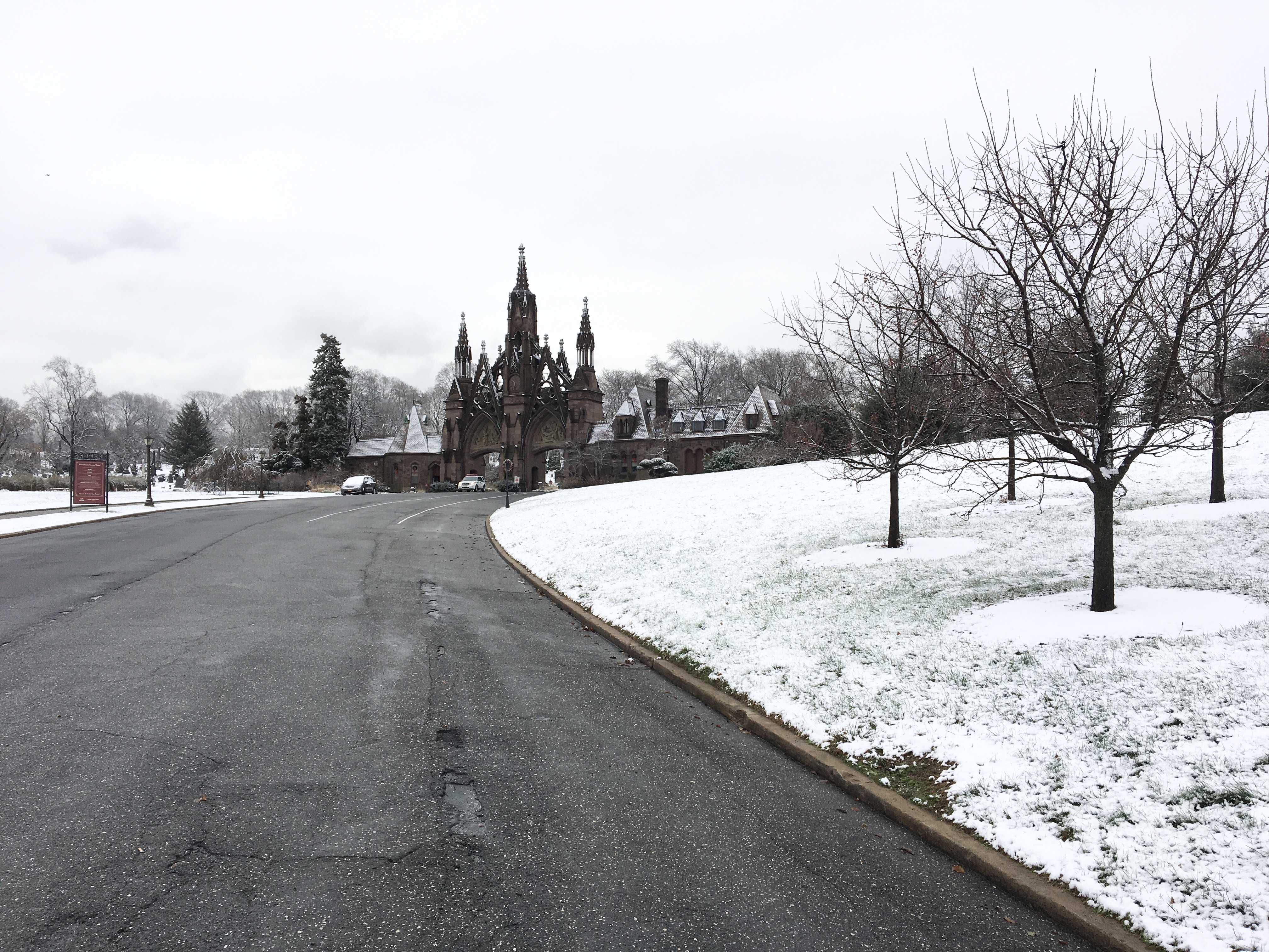 The Green-Wood Cemetery main gates seen from the bottom of a long and curving driveway. On either side of the driveway is the cemetery's grassy landscape covered in fresh white snow; barren trees are in the foreground on the right while evergreens tower in the distance behind the Gothic Revival, brownstone gates. The sky is a hazy grey with snow clouds lingering. 