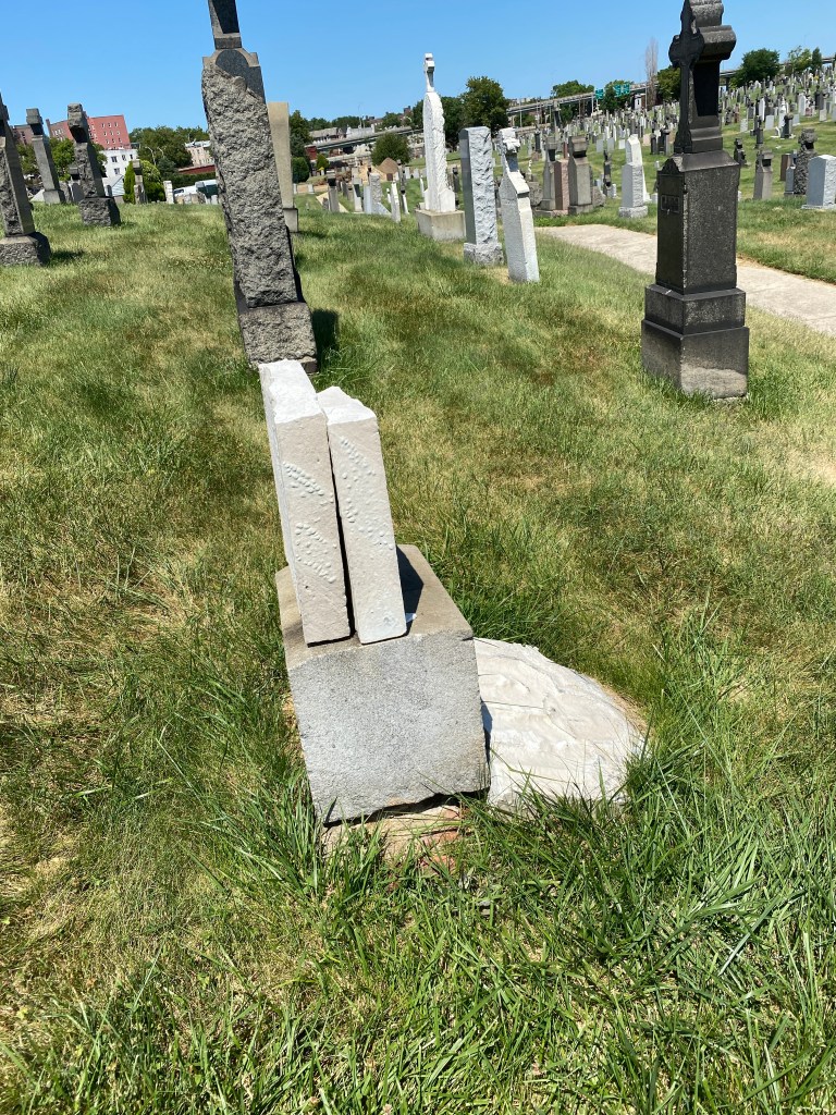 Side view of a white, stone, tablet headstone broken into three pieces. The top piece is laying flat on the ground while the middle piece leans against the bottom, which is seated on a base.