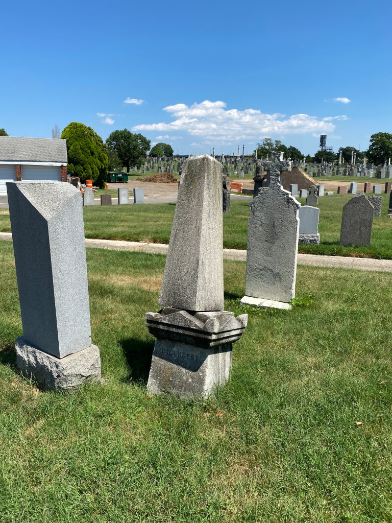 A stone obelisk burial marker is standing next to other markers. The obelisk is attached to a base but the column is leaning excessively to the back and side of the base.
