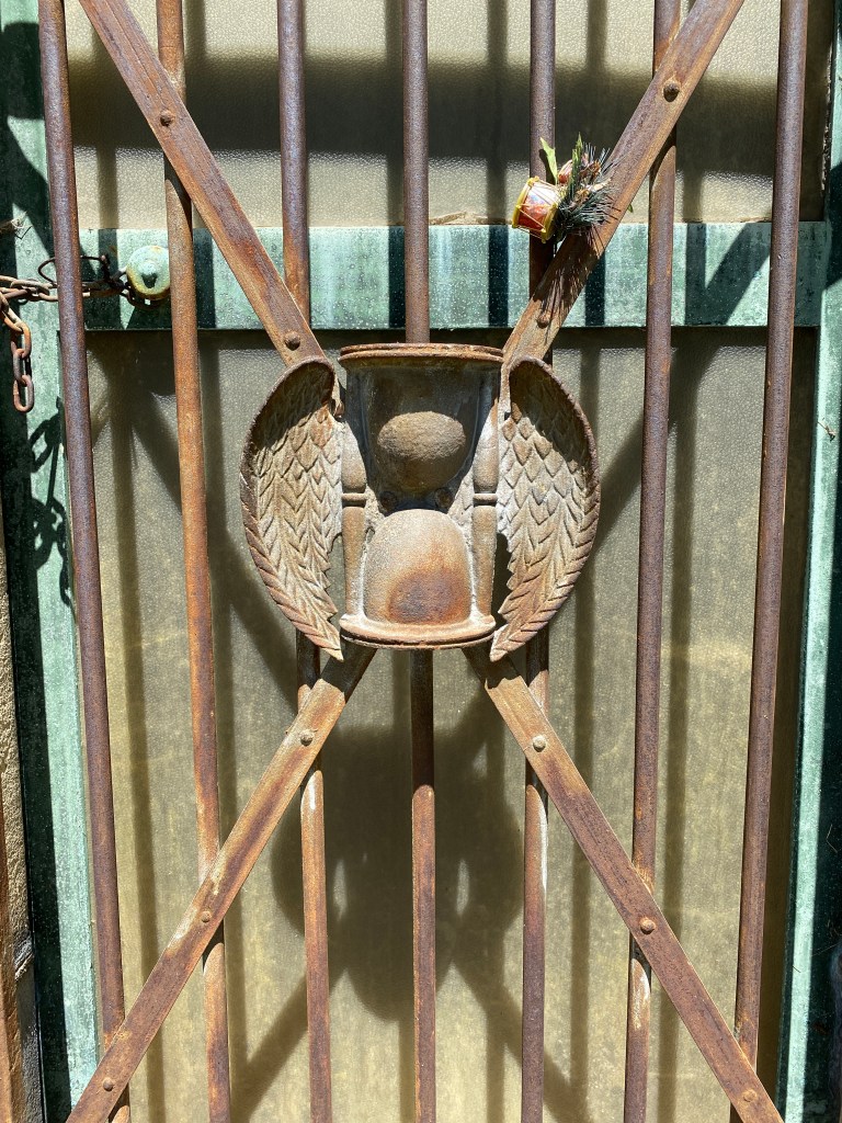 A close up picture of an rusty metal gate to a mausoleum. The gate is not solid, but rather made of seven vertical metal rod. Two, diagonal, metal bars form an "X" across the gate; atop the "X" is mounted a large rusted feather-winged hourglass.